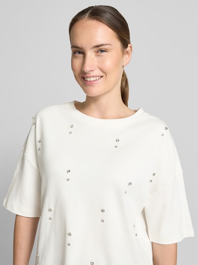 Frauenmodel trägt T-Shirt aus reiner Baumwolle von Christian Berg Woman in Weiß mit Ziersteinen, lockere Passform, Nahaufnahme Oberkörper.