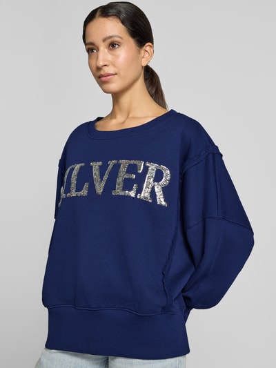 Frauenmodel trägt Oversized Sweatshirt aus reiner Baumwolle Modell 'Wide Silver' von miss goodlife in Dunkelblau mit silbernem Statement-Print, Ziernähte und elastische Abschlüsse, Nahaufnahme Oberkörper.