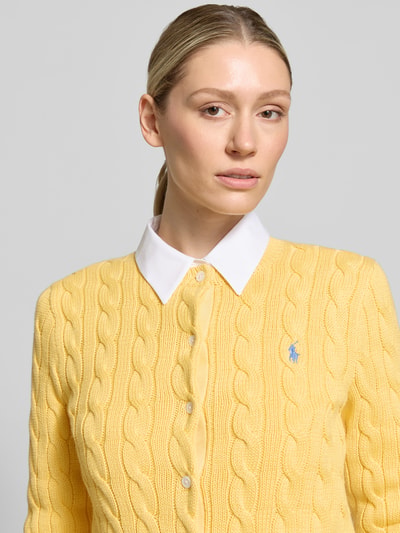 Frauenmodel trägt Regular Fit Strickjacke aus reiner Baumwolle von Polo Ralph Lauren in Optik: Unifarben, Zopfstrick, Logo-Stitching, einreihige Knopfleiste, Rundhals, Langarm, Ärmelabschluss: Gerippt, Materialverarbeitung: Zopfstrick, Farbe gelb, Struktur betont, Nahaufnahme Oberkörper.