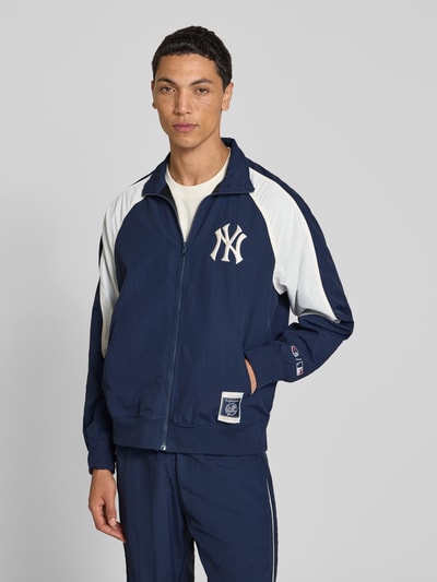 Männermodel trägt Relaxed Fit Tracksuit mit Logo-Patch von Champion in Dunkelblau mit weißen Details, Reißverschluss und Stehkragen, Nahaufnahme Oberkörper. Rückenlänge 68 cm, Bundweite 39 cm, elastische Abschlüsse.