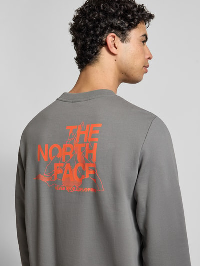 Męski model nosi Bluza z nadrukiem z logo Sweatshirt mit Label-Print od The North Face, bawełna, krój regular fit, prążkowane wykończenia, okrągły dekolt, długość tyłu 69 cm, długość rękawów 72 cm, szara z pomarańczowym nadrukiem, zdjęcie z tyłu, detal.