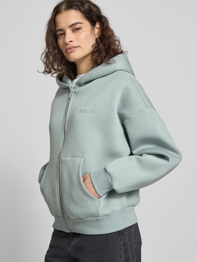 Frauenmodel trägt Sweatjacke mit Logo und Känguru-Tasche von Review, hellgrün, Baumwoll-Mix, Kapuze, Relaxed Fit, Reißverschluss, Ganzkörperansicht von vorne