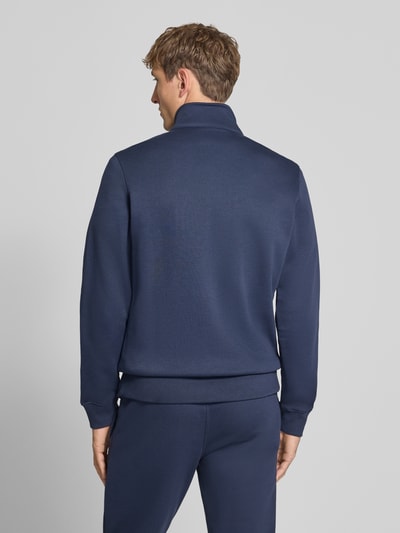 Męski model nosi Sweatanzug mit Label-Details od Champion, mieszanka bawełny, prosty krój, stójka, zamek błyskawiczny, wpuszczane kieszenie boczne, elastyczny pas, kolor granatowy, tekstura gładka, widoczne detale z logo, Szerokość w rozmiarze M: 76,42 cm, Długość tyłu w rozmiarze M: 70 cm, Szerokość ramion w rozmiarze M: 51 cm, Długość rękawów w rozmiarze M: 69 cm, Długość wewnętrzna nogawki w rozmiarze M: 76 cm, widok całej sylwetki z tyłu.
