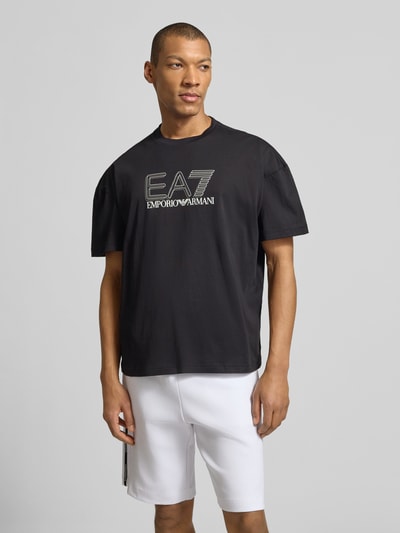 Męski model trägt T-shirt mit Logo und Rundhalsausschnitt von EA7 Emporio Armani, czarna bawełna, krój regular fit, logo, okrągły dekolt, długość tyłu w rozmiarze M: 68 cm, Ganzkörperansicht von vorne.