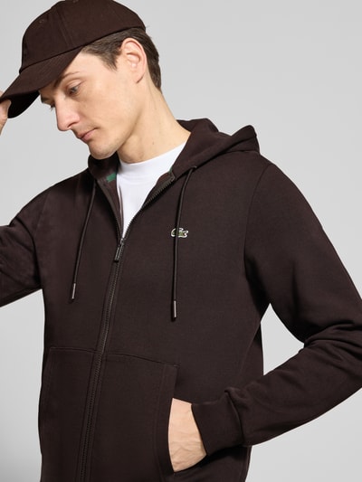 Męski model nosi Regular Fit Sweatjacke aus Baumwoll-Mix od Lacoste, jednokolorowa, krój regular fit, zamek błyskawiczny, naszywka z logo, kaptur z tunelem, długi rękaw, prążkowane zakończenia, przednie kieszenie, dzianina dresowa, mieszanka bawełny, ciemny brąz, gładka tekstura. Ujęcie: z przodu, zbliżenie na górną część ciała.