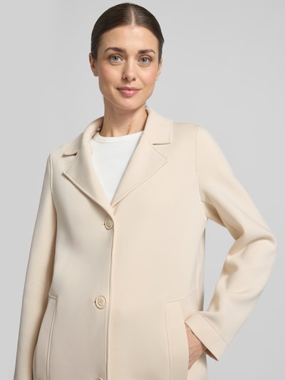 Vrouwenmodel draagt Regular Fit Kurzmantel mit Kapuze van s.Oliver RED LABEL in effen lichtbeige scuba, reverskraag, afneembare capuchon, enrijige knopen, lange mouwen, zij-naad steekzakken, middeleeuws textuur, detailopname bovenlichaam.