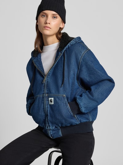 Frauenmodel trägt Jeansjacke mit Kapuze und Reißverschluss von Carhartt Work In Progress in Blau, strukturierte Baumwolle, Regular Fit, Nahaufnahme Oberkörper.
