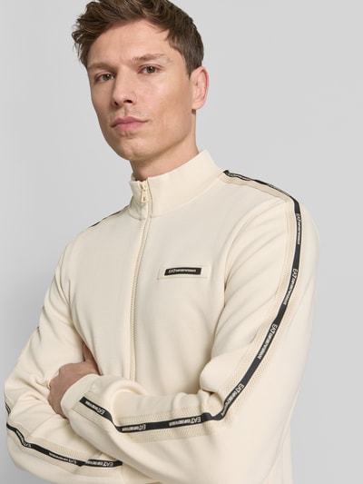 Männermodel trägt Sweatjacke mit Stehkragen und Zweiwege-Reißverschluss von EA7 Emporio Armani in Creme mit Label-Detail, Nahaufnahme Oberkörper. Rückenlänge 68 cm, Ärmellänge 67 cm, Regular Fit, Eingrifftaschen.