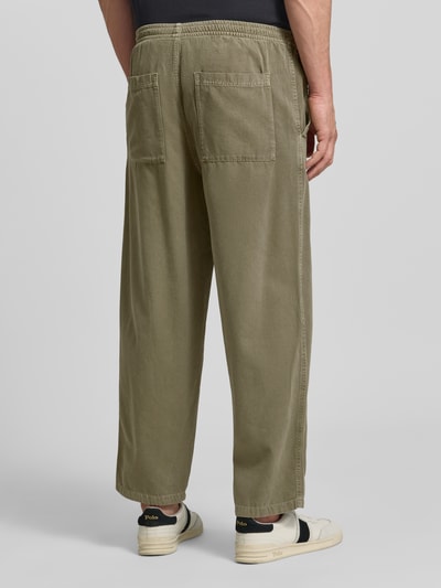 Männermodel trägt Jog Pants mit französischen Eingrifftaschen Modell 'LYCAZ' von American Vintage, reine Baumwolle, gerade Passform, olivgrün, elastischer Bund, Gesäßtaschen, Ganzkörperansicht Rückansicht.