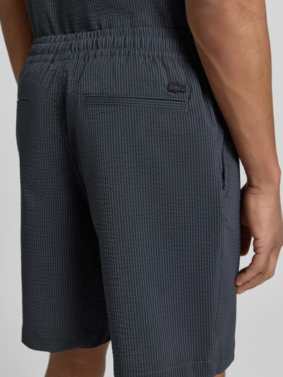 Männermodel trägt Loose Fit Shorts aus Baumwoll-Mix von Lacoste, Optik:Allover-Muster, Gestreift, Label-Stitching-Applikation, Bundabschluss: Elastisch mit Tunnelzug, Taschen: Seitliche Eingrifftaschen, Materialverarbeitung: Seersucker, Passform: Regular-Fit, Farbe dunkel, Textur fein strukturiert, Ganzkörperansicht Rückansicht.