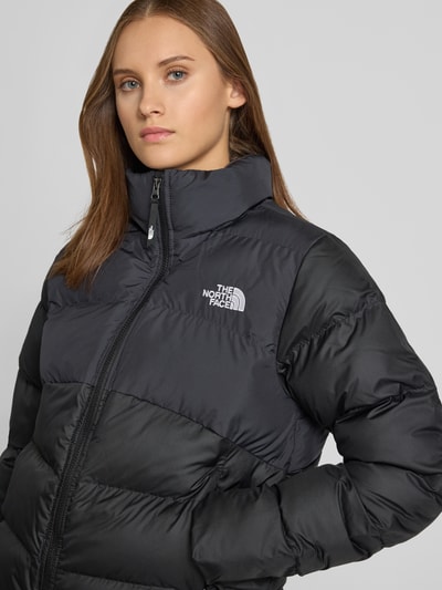 Kobiece model prezentuje czarno-szarą kurtkę pikowaną z wyhaftowanym logo, zamkiem błyskawicznym i kieszeniami zapinanymi na zamek błyskawiczny, Model 'SAIKURU' od The North Face, krój regular fit, stójka, szerokość w rozmiarze S: 43 cm, szerokość klatki piersiowej w rozmiarze S: 53 cm, długość tyłu w rozmiarze S: 59 cm, długość rękawów w rozmiarze S: 62 cm, zbliżenie na górną część ciała