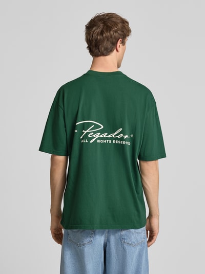 Mężczyznamodel nosi Oversized T-Shirt aus reiner Baumwolle Modell 'Pike' von Pegador, wyhaftowane logo, okrągły dekolt, krój oversized, długość tyłu 71 cm, zielony kolor, gładka faktura, Ganzkörperansicht Rückansicht.