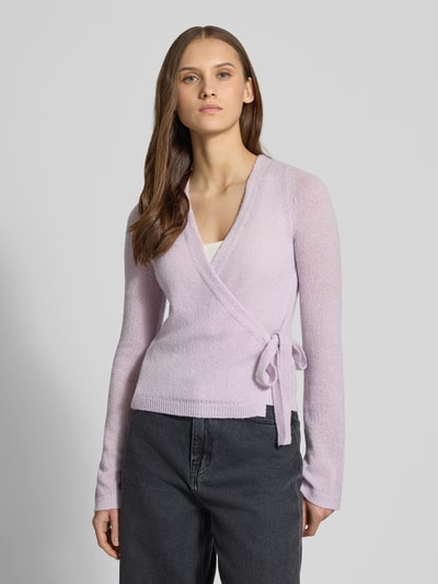 Frauenmodel trägt Strick-Cardigan in Wickeloptik von Review in Flieder, V-Ausschnitt, lange Ärmel, Schleifenverschluss, Ganzkörperansicht von vorne.