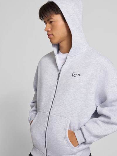 Männermodel trägt Sweatjacke mit Label-Stitching und Känguru-Tasche von Karl Kani, hellgrau, Baumwoll-Mix, Ganzkörperansicht von vorne.