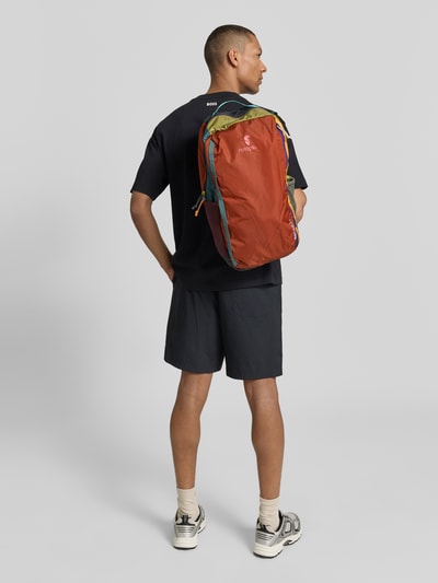 Männermodel trägt Rucksack mit Logo-Print Modell 'Batac' von Cotopaxi in Rot, Grün und Blau, Hauptfach mit Reißverschluss, Ganzkörperansicht Rückansicht.