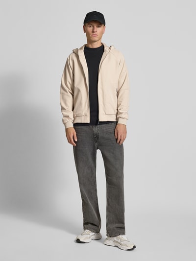 Männermodel trägt Jacke mit Kapuze Modell 'JAXON' von Jack & Jones, materiał z domieszką stretchu, luźny krój, jasny kolor, zamek, kieszenie, ściągacze, długość tyłu w rozmiarze M 66 cm, szerokość ramion w rozmiarze M 41 cm, szerokość klatki piersiowej w rozmiarze M 56 cm, długość rękawa w rozmiarze M 74 cm, całopostaciowy widok z przodu
