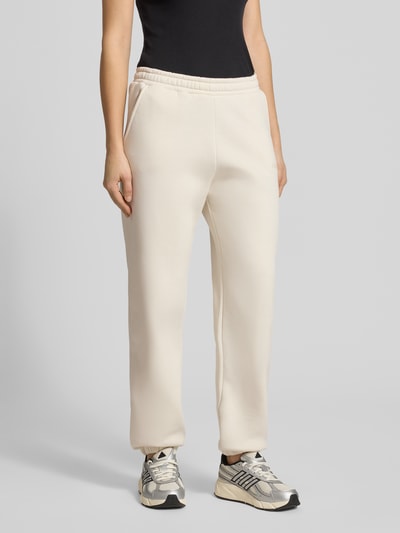 Frauenmodel trägt Basic Sweatpant mit Bündchen am Saum und Seitentaschen von Review, cremefarben, Baumwoll-Mix, lockere Passform, Label-Print, Logo-Stickerei, elastische Abschlüsse, Seitentaschen, Ganzkörperansicht von vorne.