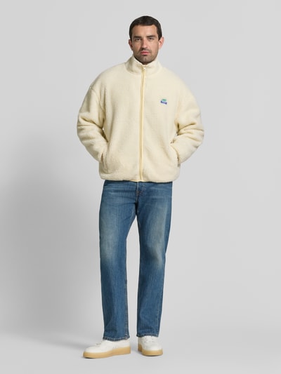 Männermodel trägt fleecejack met opstaande kraag en labeldetail model 'HOKTOWN' van American Vintage in ecru, relaxed fit, steekzakken, ritssluiting, ruglengte maat M/L 69 cm, schouderbreedte 56 cm, borstwijdte 48 cm, mouwlengte 55 cm, ganzkörperansicht van voren.