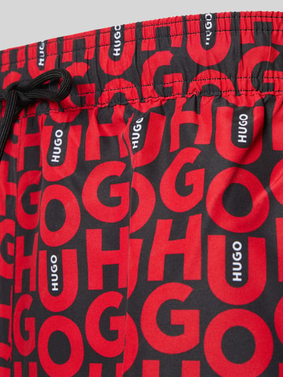 Produktfoto, Badeshorts mit Allover-Muster Modell 'NALU' von HUGO, rote und schwarze Farben, Label-Schriftzug, elastischer Bund, seitliche Taschen