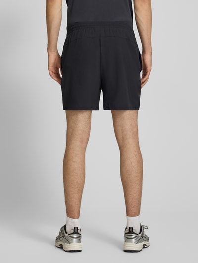 Mężczyzna nosi szorty z elastycznym pasem i logo od New Balance, krój regular fit, tunel, szerokość w rozmiarze M: 39 cm, długość wewnętrzna nogawki: 12 cm, czarny kolor, gładka tekstura, pełny widok od tyłu.