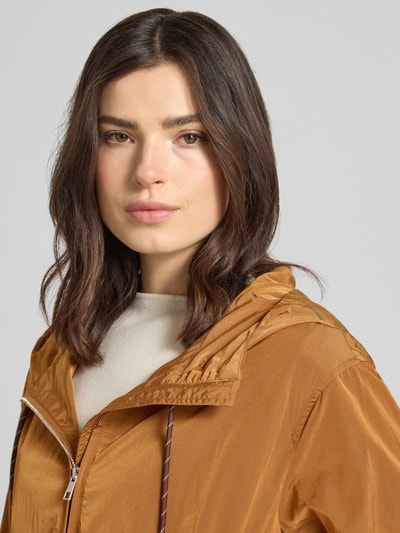 Kobiece model nosi Trekking Jacke mit Kapuze und Reißverschlusstaschen od Rich & Royal w kolorze miodowym, tekstura lekko połyskująca, prosty krój, kaptur z trokiem, zamek błyskawiczny, kieszenie zapinane na zamek błyskawiczny, elastyczne wykończenia, długość tyłu w rozmiarze 36: 58 cm, szerokość ramion w rozmiarze 36: 48 cm, szerokość klatki piersiowej w rozmiarze 36: 56 cm, długość rękawów w rozmiarze 36: 59 cm, zbliżenie na górną część ciała.