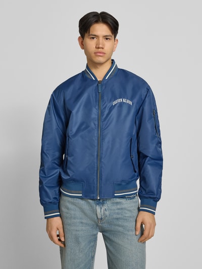 Männermodel trägt Regular Fit College-Jacke mit Label Stitching van Calvin Klein Jeans in look: effen blauw met contraststrepen, lange mouw, college-kraag, ritssluiting, applicatie met labelstitching, geribde mouwboord en rompzoom, klepzakken, regular fit pasvorm, voorkant, ganzkörperansicht van voren.