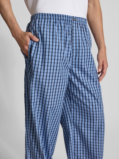 Mężczyzna nosi piżamowe spodnie w niebieską kratę, piżama-Hose aus reiner Baumwolle von VETEMENTS, krój relaxed fit, elastyczny pas, wpuszczane kieszenie, detal z logo, długość wewnętrzna nogawki w rozmiarze M: 86 cm, szerokość w rozmiarze M: 46 cm, detal: zbliżenie na nogawkę i kieszeń, perspektywa: zbliżenie na górną część spodni.