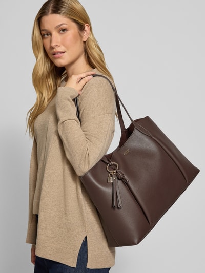 Frauenmodel trägt Tote Bag in Leder-Optik Modell 'MELODY' von GUESS in Dunkelbraun mit Logo-Detail, Ganzkörperansicht von vorne.