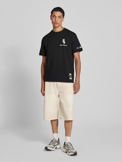 Męski model nosi Relaxed Fit T-Shirt aus reiner Baumwolle od Champion, czarny z nadrukiem i wyhaftowanym logo, bawełna, krój relaxed fit, długość tyłu w rozmiarze M: 70 cm, widok całej sylwetki z przodu.