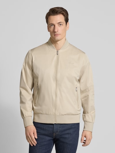 Männermodel trägt Bomberjacke mit Reißverschlusstaschen Modell 'MATTE' von CK Calvin Klein in Beige, glatte Struktur, Ganzkörperansicht von vorne.