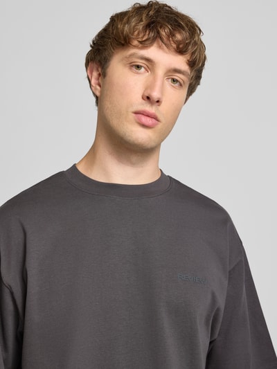 Męski model nosi Oversized T-Shirt mit Logo-Applikation od REVIEW, bawełna, krój oversized fit, okrągły dekolt, długość tyłu 70 cm, rozmiar M, kolor grafitowy, prosty materiał, gładka tekstura. Zbliżenie na górną część ciała z przodu.
