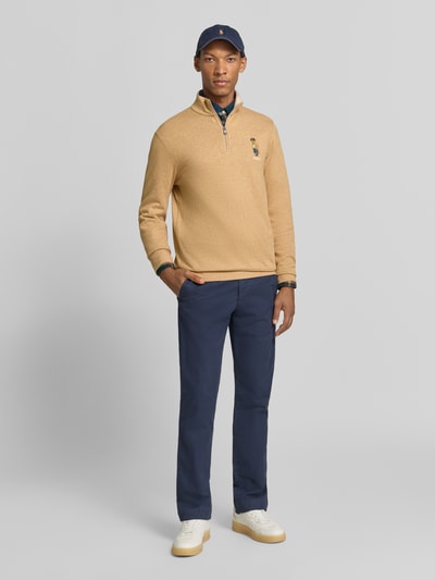 Męski model nosi Regular Fit Pullover aus Baumwoll-Mix mit Viskose-Anteil mit Polo-Bear-Stitching von Polo Ralph Lauren, beżowy sweter, granatowe spodnie, białe buty, granatowa czapka, tło jasnoszare, Herren Pullover von Polo Ralph Lauren, całopostaciowy widok z przodu.