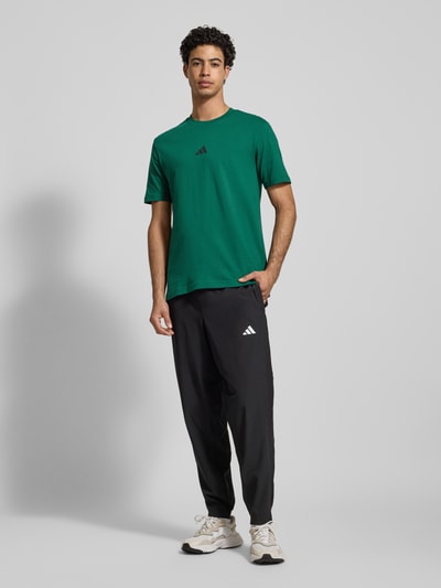 Männermodel trägt T-Shirt mit Label-Print von adidas Sportswear in Grün aus reiner Baumwolle, Regular-Fit, gerippter Rundhalsausschnitt, Ganzkörperansicht von vorne.