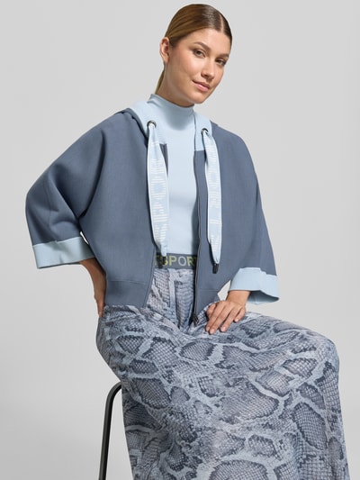 Frauenmodel trägt Relaxed Fit Sweatjacke aus Viskose-Mix mit Logo-Detail von Marc Cain in Blau, seitliche Nahaufnahme Oberkörper, Damen Sweatjacke, Kapuze mit Tunnelzug, Reißverschluss, elastischer Bund, Rückenlänge 51 cm, Ärmellänge 54 cm, glatte Struktur.