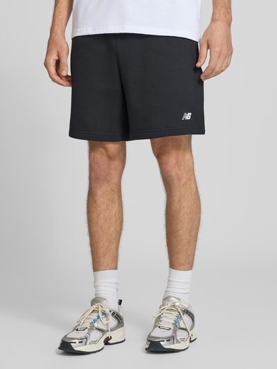 Mężczyzna nosi Relaxed Fit Shorts mit Logo-Stitching od New Balance, czarne, mieszanka bawełny, wyhaftowane logo, elastyczny pas z tunelem, Szerokość w rozmiarze M: 42 cm, Długość wewnętrzna nogawki: 18 cm, zdjęcie całej sylwetki od przodu.