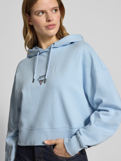 Vrouwenmodel draagt Relaxed Fit Hoodie mit Logo-Stitching Modell 'Baker' van Guess Jeans in lichtblauw, katoen, kap met trekkoord, elastische boorden, logo op de borst, Nahaufnahme bovenlichaam.