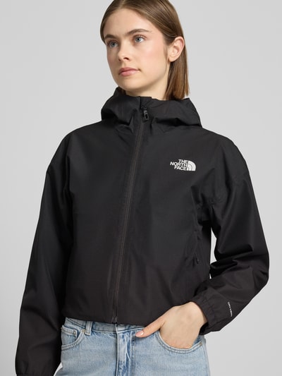 Modelka kobieta ma na sobie Cropped Jacke mit Logo und Reißverschluss od The North Face, czarna tkanina, logo, kaptur, widok na górną część ciała z przodu, długość tyłu w rozmiarze S: 58 cm, szerokość ramion w rozmiarze S: 54 cm, szerokość klatki piersiowej w rozmiarze S: 50 cm, długość rękawa w rozmiarze S: 56 cm