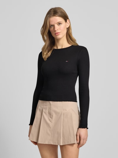 Modelka nosi Slim Fit Langarmshirt aus Baumwoll-Mix von Tommy Jeans, czarny kolor, detal z logo, okrągły dekolt, długi rękaw, mieszanka bawełny, drobne prążkowanie, zakończenie frill, wzór w paski, widok całej sylwetki od przodu.