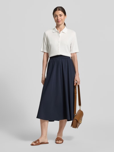 Frauenmodel trägt Regular Fit Poloshirt mit durchgehender Knopfleiste von OPUS, Viskose-Mix mit Stretch-Anteil, Ganzkörperansicht von vorne. Rückenlänge bei Größe 36: 54 cm, Farbe Weiß, kombiniert mit dunkelblauem Rock und braunen Accessoires.
