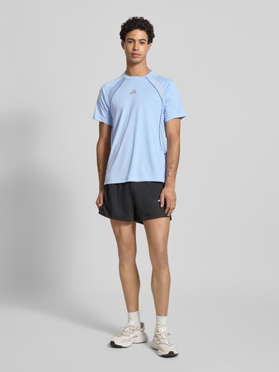 Männermodel trägt T-Shirt mit Label-Print von adidas Training in Hellblau, Regular Fit, Rundhalsausschnitt, schwarze Shorts, Ganzkörperansicht von vorne.