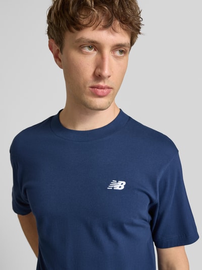 Männermodel trägt T-Shirt mit Logo und Rundhalsausschnitt von New Balance in Dunkelblau, Reine Baumwolle, Regular-Fit, Nahaufnahme Oberkörper. Rückenlänge bei Größe M: 74 cm.