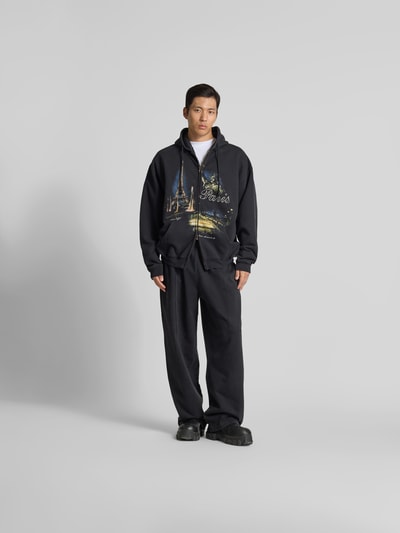Männermodel trägt Oversized Sweatjacke mit Foto-Print von VETEMENTS aus Baumwoll-Mix mit Zweiwege-Reißverschluss, Kapuze mit Tunnelzug, dunkle Farben, Rückenlänge bei Größe M 74 cm, Schulterbreite 68 cm, Ärmellänge 67 cm, Oversized-Fit, Ganzkörperansicht von vorne.