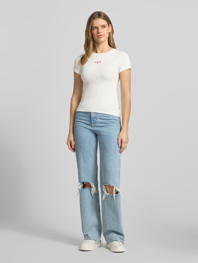 Vrouwenmodel draagt T-Shirt in Ripp-Optik mit Logo van Guess Jeans, wit, slim fit, katoen-elastaan mix, ronde hals, geribbelde structuur, logo-opdruk, ruglengte maat S 54 cm, full body voorkant.