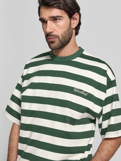 Männermodel trägt Relaxed Fit T-Shirt mit Logo-Stitching Modell 'Beam' von Woodbird aus reine Baumwolle, grüne und weiße Streifen, Rundhalsausschnitt, Nahaufnahme Oberkörper.