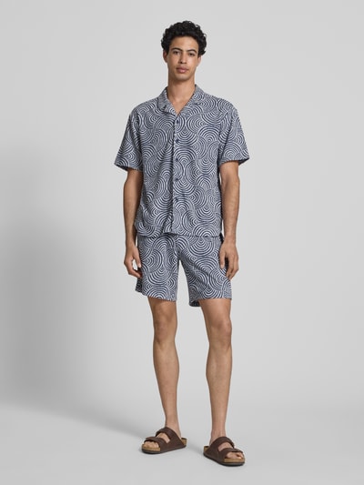 Męski model nosi Relaxed Fit Freizeithemd mit Baumwoll-Anteil Modell 'CLEMENT' od Only & Sons, materiał frotte, wzór allover, koszula z krótkim rękawem, resortowy kołnierzyk, niebiesko-białe okręgi, widok całej sylwetki z przodu.