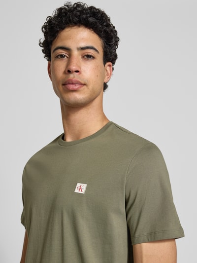 Mannelijk model draagt Regular Fit T-Shirt aus reiner Baumwolle van Calvin Klein Jeans in effen olijfgroen, label patch, rondhals, korte mouw, puur katoen. Nahaufnahme bovenlichaam.