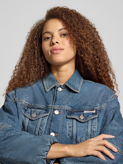 Frauenmodel trägt Cropped Jeansjacke mit Knopfleiste von Calvin Klein Jeans in Blau, Reine Baumwolle, Lockerer-Schnitt, Brusttaschen, Nahaufnahme Oberkörper.