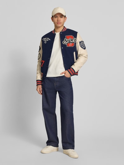 Męskimodel nosi College-Jacke mit Patches und Eingrifftaschen von Superdry, mieszanka bawełny, krój regular fit, stójka, listwa guzikowa, wpuszczane kieszenie, długość tyłu w rozmiarze M: 65 cm, szerokość ramion w rozmiarze M: 45 cm, szerokość klatki piersiowej w rozmiarze M: 49 cm, długość rękawów w rozmiarze M: 70 cm, widok całej sylwetki z przodu
