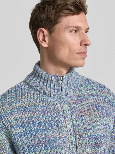Männermodel trägt Relaxed Fit Strickjacke aus Baumwoll-Woll-Mix mit Viskose-Anteil Modell 'UN-WASTED' von Armedangels, mehrfarbig, Strickoptik, Reißverschluss, Logo-Detail, Stehkragen, langarm, gerippte Abschlüsse, Nahaufnahme Oberkörper.