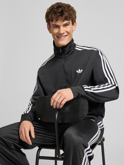 Männermodel trägt Sweatjacke mit Reißverschluss von adidas Originals in Schwarz mit weißen Streifen und Label-Stitching, Nahaufnahme Oberkörper.